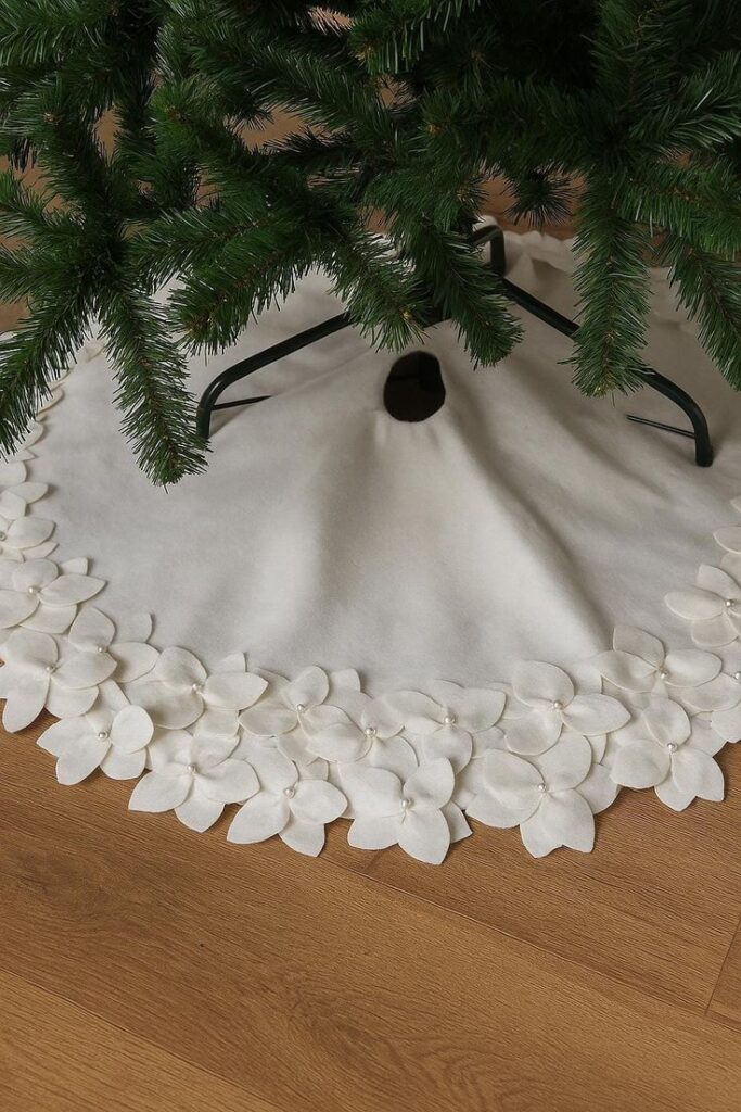 White felt Christmas tree skirt decorated with floral appliques, elegantly placed on a wooden floor beneath a green tree.