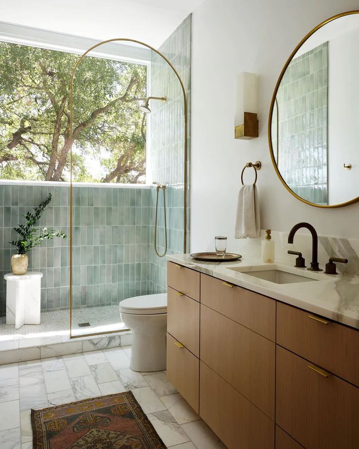 Une salle de bain apaisante Modern bathroom with green tiled shower, round mirror, wooden vanity, and large window overlooking trees, creating a serene, natural ambiance. | Sky Rye Design Modern bathroom with green tiled shower, round mirror, wooden vanity, and large window overlooking trees, creating a serene, natural ambiance.