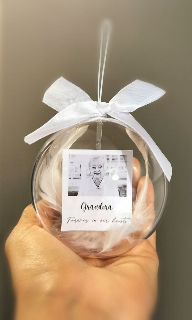 Unique Personalised Christmas Baubles with Photos Engravings and More Memorial ornament with a photo of a grandma, adorned with a white bow and feathers, held in a hand. | Sky Rye Design Memorial ornament with a photo of a grandma, adorned with a white bow and feathers, held in a hand.