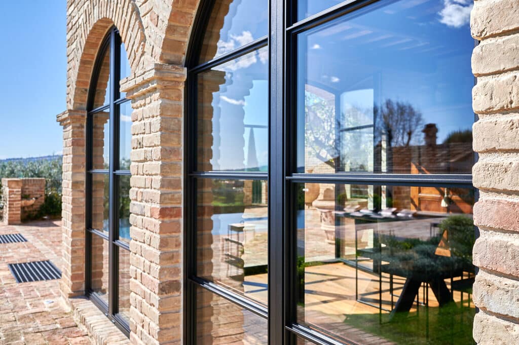 Modern arched windows reflect a sunny outdoor view, framed by brick walls, creating a seamless connection between indoor and outdoor spaces.