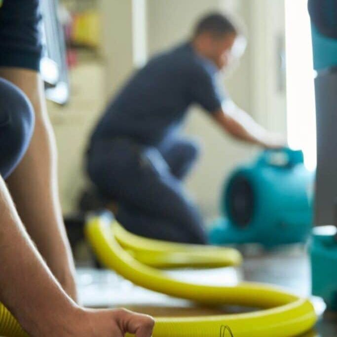 Water damage restoration experts using equipment to dry a wooden floor. Yellow hose and blue air mover visible in the background.