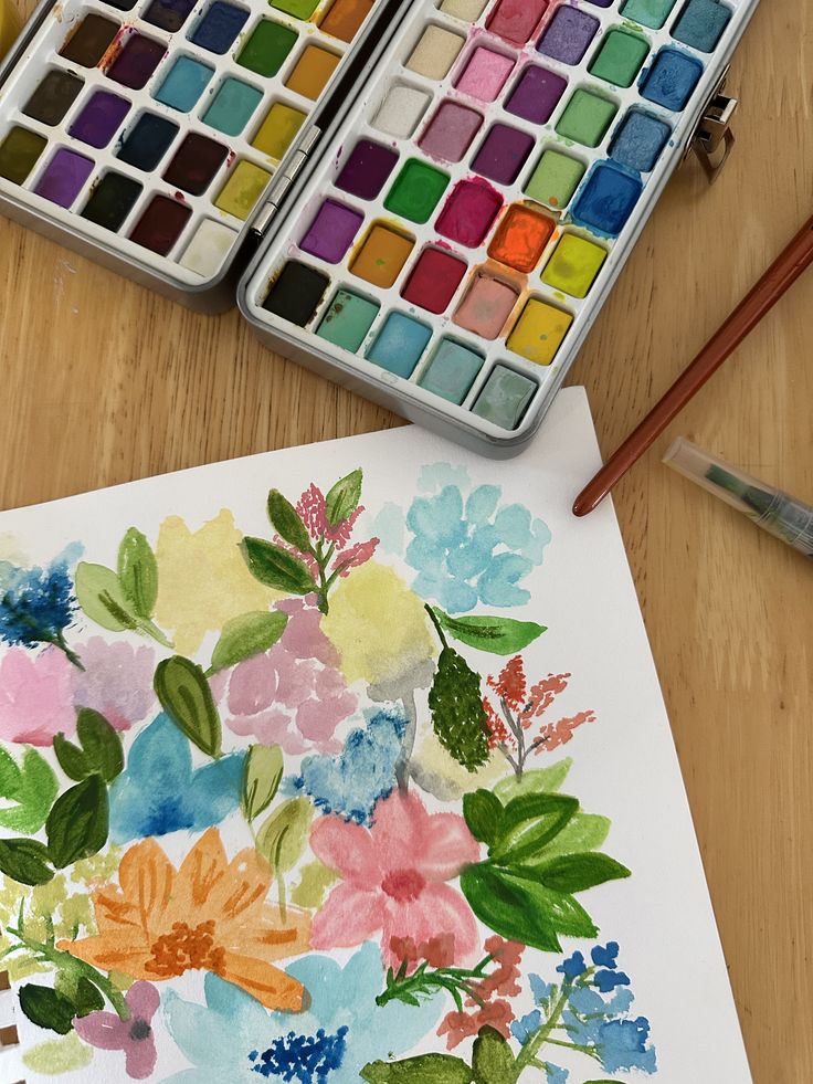 Watercolor Colorful watercolor palette and floral painting on a wooden table, showcasing vibrant pigments and an artistic floral design. | Sky Rye Design Colorful watercolor palette and floral painting on a wooden table, showcasing vibrant pigments and an artistic floral design.