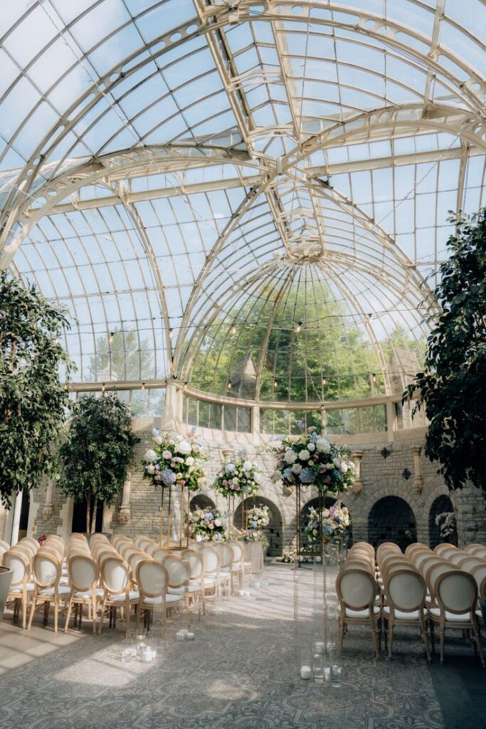 Nesreen and Tom - Permitted to post Elegant glass-roofed wedding venue with floral arrangements, white chairs, and stone walls, creating a romantic and sophisticated atmosphere. | Sky Rye Design Elegant glass-roofed wedding venue with floral arrangements, white chairs, and stone walls, creating a romantic and sophisticated atmosphere.