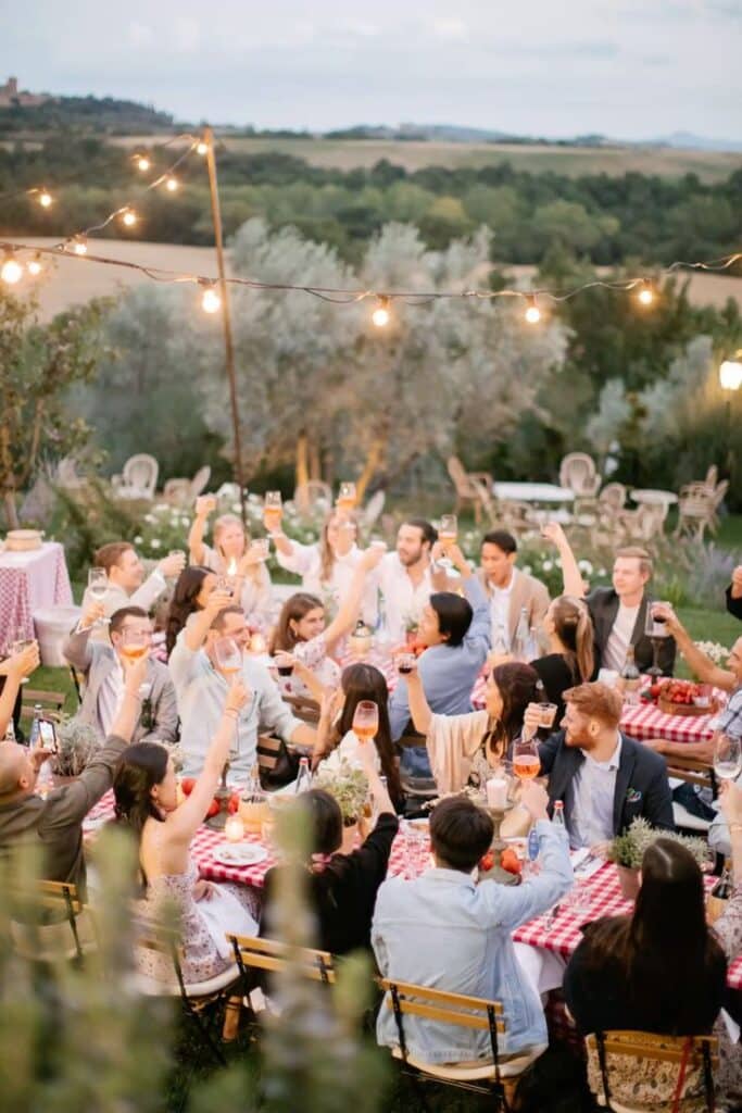 Wedding pizza party in Tuscany Outdoor celebration with friends raising glasses at a table under string lights in a scenic countryside setting with greenery. | Sky Rye Design Outdoor celebration with friends raising glasses at a table under string lights in a scenic countryside setting with greenery.