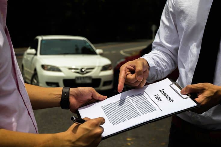 Two people discuss an insurance policy with a white car in the background, highlighting accident coverage and claims negotiation.