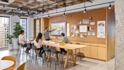 Modern office meeting with four people collaborating, using a large screen and pinned notes for creative discussion in a bright workspace.