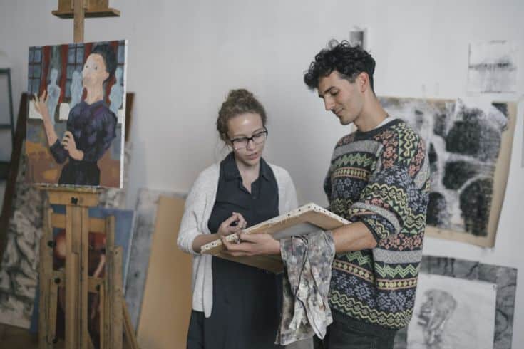 Two artists discussing a painting in a studio, surrounded by artworks and canvases. Creative collaboration and artistic process.