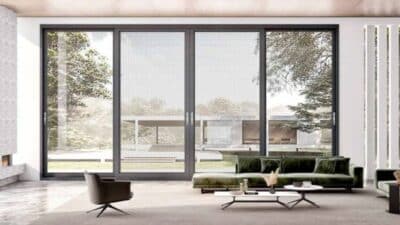 Modern living room with large glass doors, green sofa, and black chair, opening to a serene garden view. Bright, minimalist interior design.