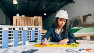 Female architect designing building models at a desk, wearing a hard hat.