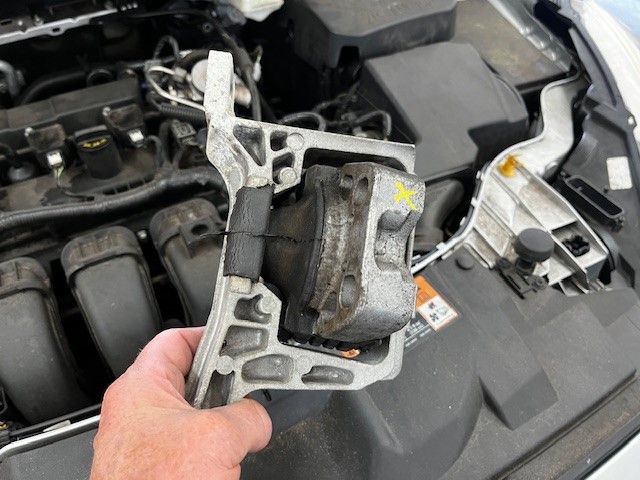Close-up of a person's hand holding a car engine mount, with the engine visible in the background, for automotive repair context.