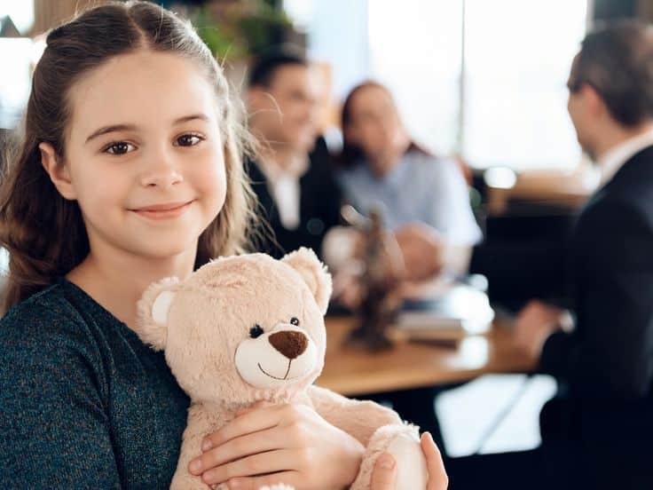 Smiling girl holding a teddy bear in a friendly office setting with blurred people in the background, conveying warmth and comfort.