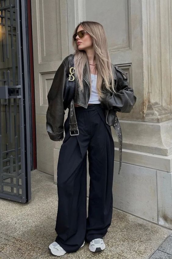 Woman in a chic leather jacket and wide-leg pants, standing stylishly with sunglasses outside a building. Fashionable street style.