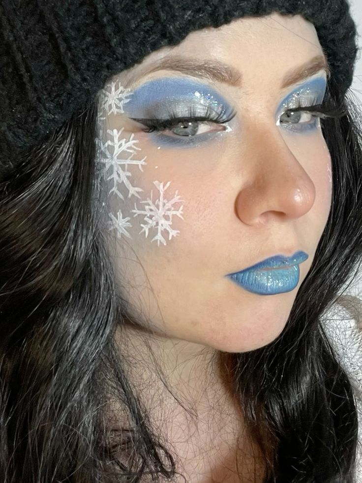 Woman with snowflake face paint, blue eye shadow, and lipstick wearing a black knit hat, showcasing winter-themed makeup artistry.