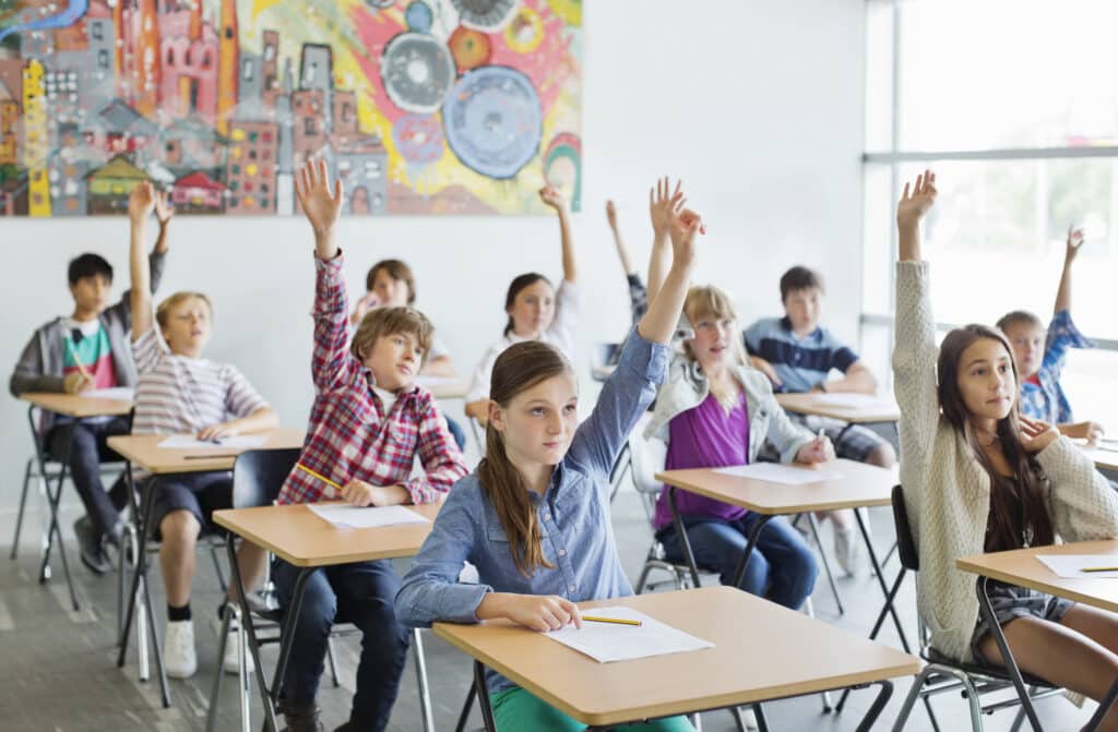 Students with arms raised in classroom Students in a classroom raising their hands to answer a question, sitting at desks in front of a colorful mural. | Sky Rye Design Students in a classroom raising their hands to answer a question, sitting at desks in front of a colorful mural.