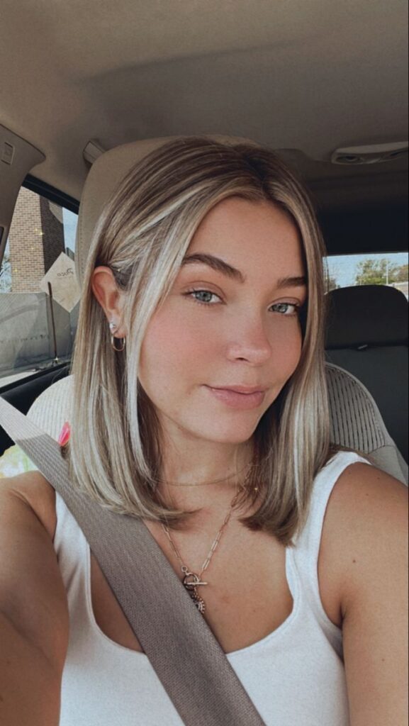 Woman with shoulder-length hair sitting in a car, wearing a white top and seatbelt, looking at the camera.