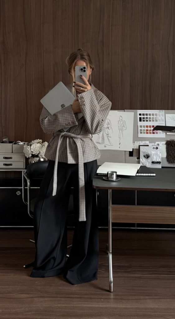 Fashionable person in stylish office with a laptop, taking a mirror selfie. Chic attire features plaid top and black wide-leg pants.