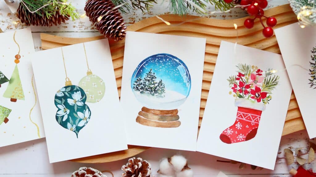 Watercolor holiday cards featuring ornaments, a snow globe, and a festive stocking surrounded by pinecones and greenery.