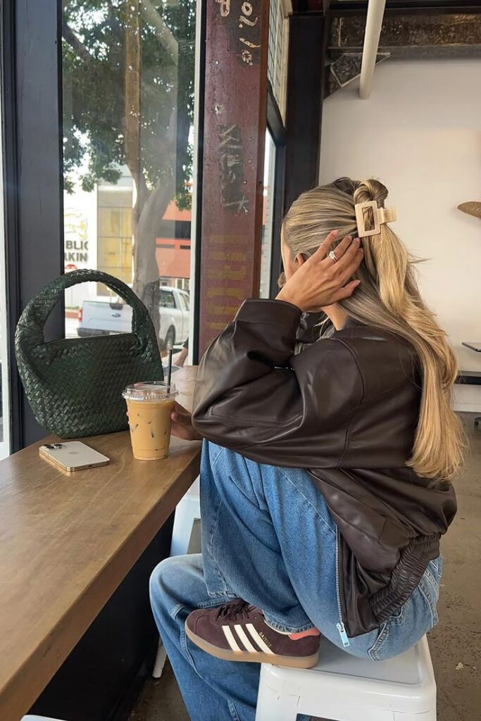 Woman in café with iced coffee, wearing leather jacket and jeans, sitting by window with phone and woven bag. Casual urban style.