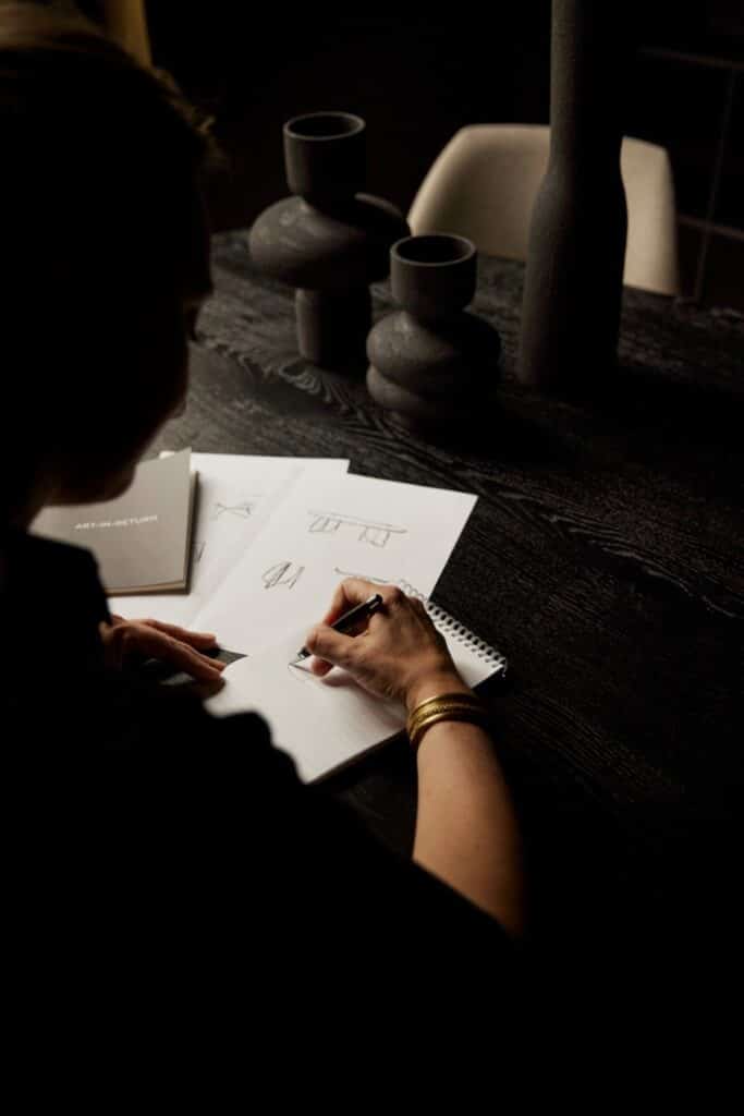 Person sketching on paper at a dark wooden table, surrounded by design sheets and modern black decor, highlighting creativity.