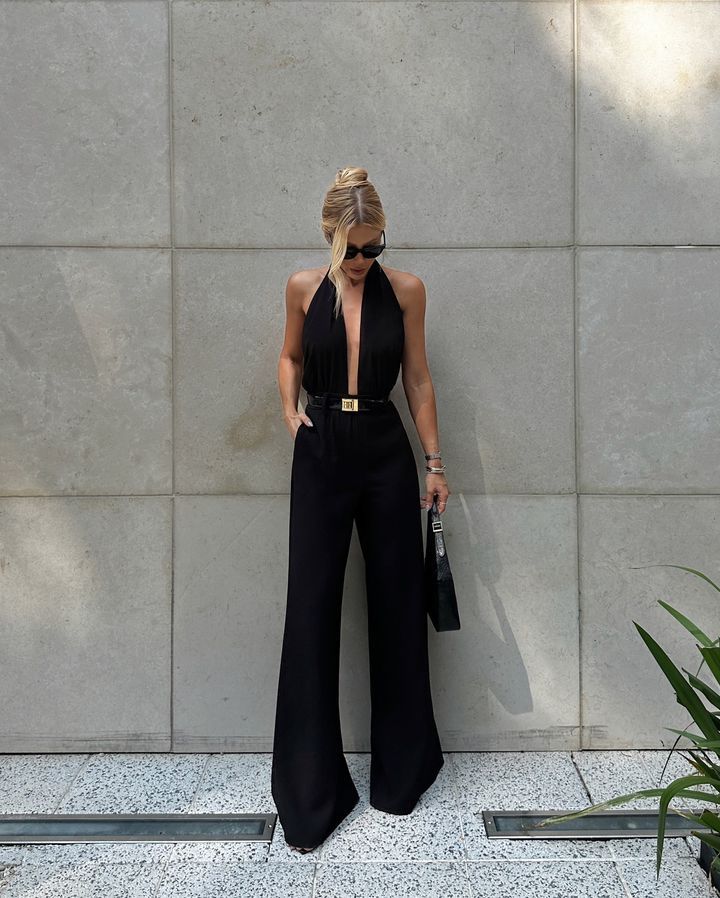 amandapaggi Fashionable woman in a black jumpsuit and sunglasses poses against a concrete wall, holding a handbag, showcasing chic style. | Sky Rye Design Fashionable woman in a black jumpsuit and sunglasses poses against a concrete wall, holding a handbag, showcasing chic style.