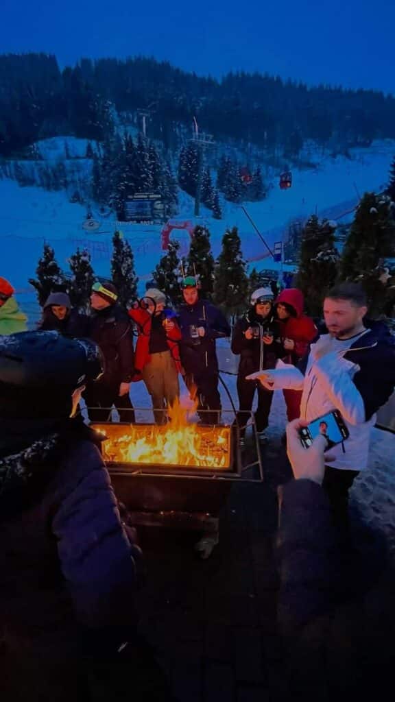 Group gathered around a warm fire on a snowy evening, enjoying wintertime outdoors with ski slopes and a cable car in the background.