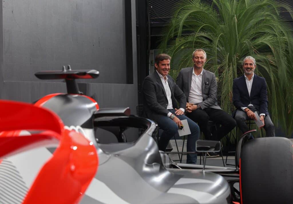 Men sit smiling behind a striking race car, with lush palm leaves in the background, capturing a moment of automotive elegance.
