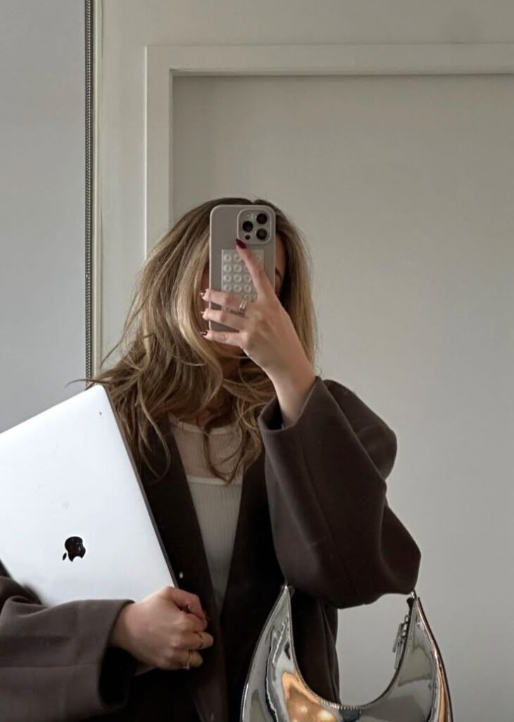 Person taking a mirror selfie holding a laptop, dressed in casual chic attire, with a stylish handbag and smartphone.