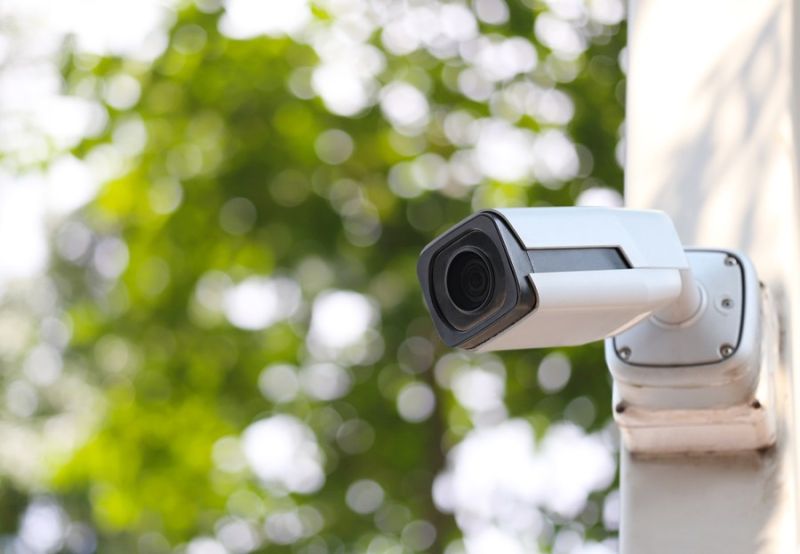 Outdoor security camera mounted on a wall, capturing green foliage in the background for enhanced surveillance coverage.