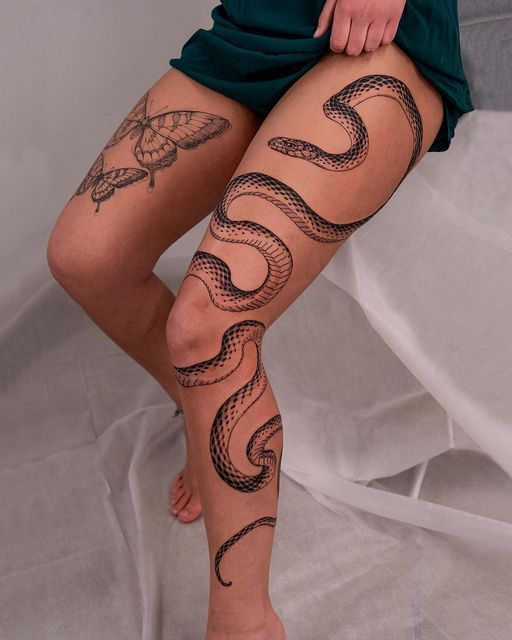 Person showcasing intricate snake and butterfly tattoos on legs, sitting against a neutral background.