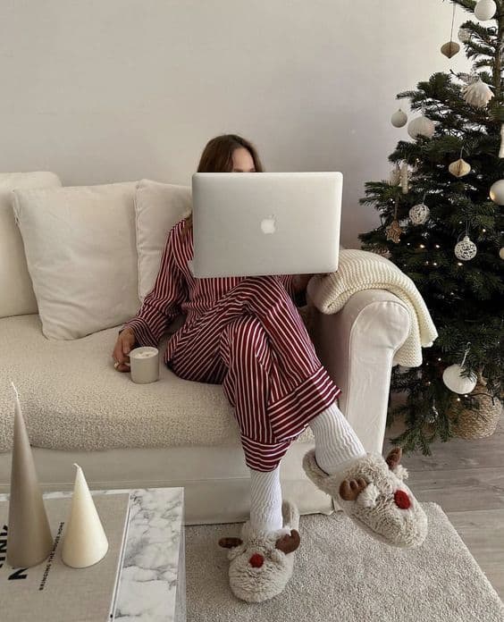 Cozy holiday scene: person in striped pajamas with laptop and mug, near Christmas tree on white couch.
