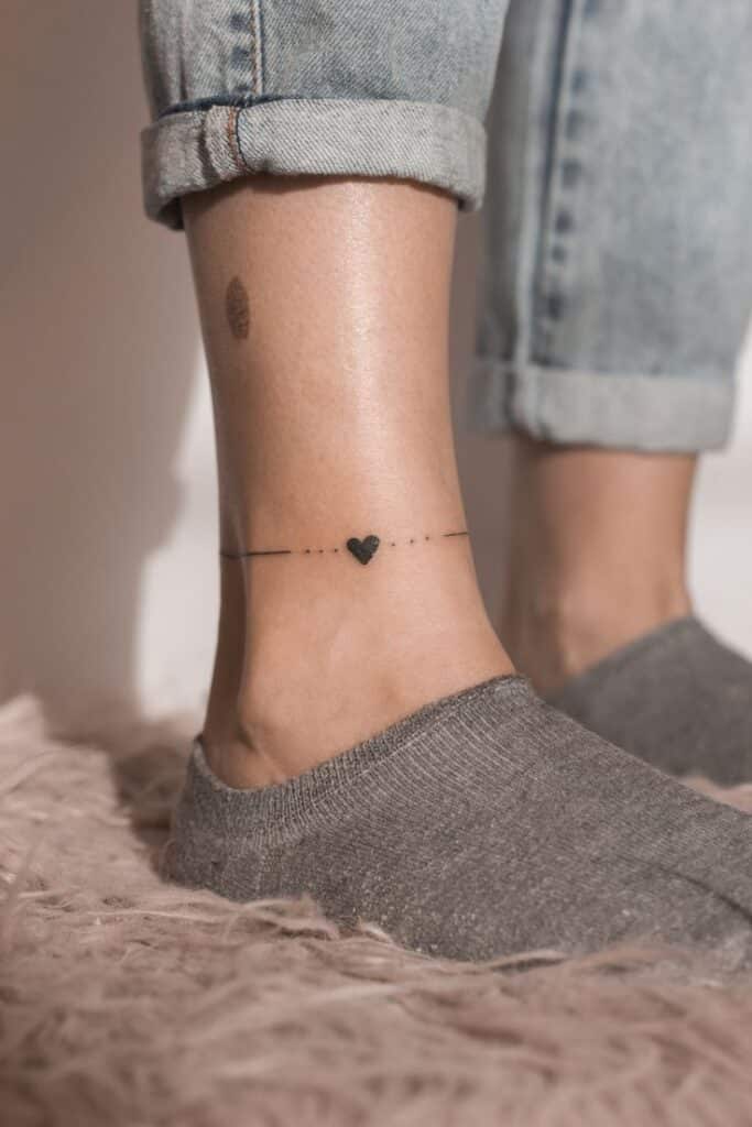 bbee2e2b18aa984a41aef189808393d6 Minimalist ankle bracelet tattoo with heart design on leg, wearing gray socks and jeans, on a soft rug. | Sky Rye Design Minimalist ankle bracelet tattoo with heart design on leg, wearing gray socks and jeans, on a soft rug.