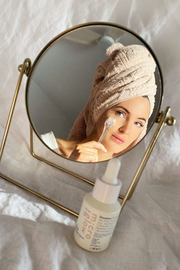 Woman in towel turban uses skincare tool in front of mirror; serum bottle on table, focusing on beauty and self-care routine.