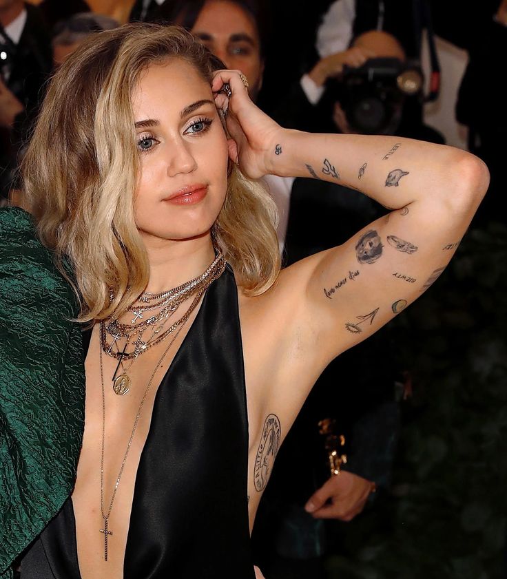 c219ff2e926c2159233b51a120b50e27 A person poses confidently on the red carpet, showcasing their tattoos and layered necklaces in a stylish black outfit. | Sky Rye Design A person poses confidently on the red carpet, showcasing their tattoos and layered necklaces in a stylish black outfit.