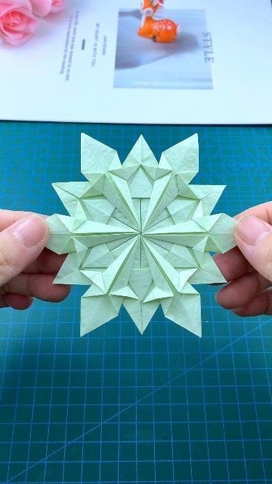 Hands hold a detailed green origami snowflake star against a grid cutting mat, showcasing intricate paper folding art.