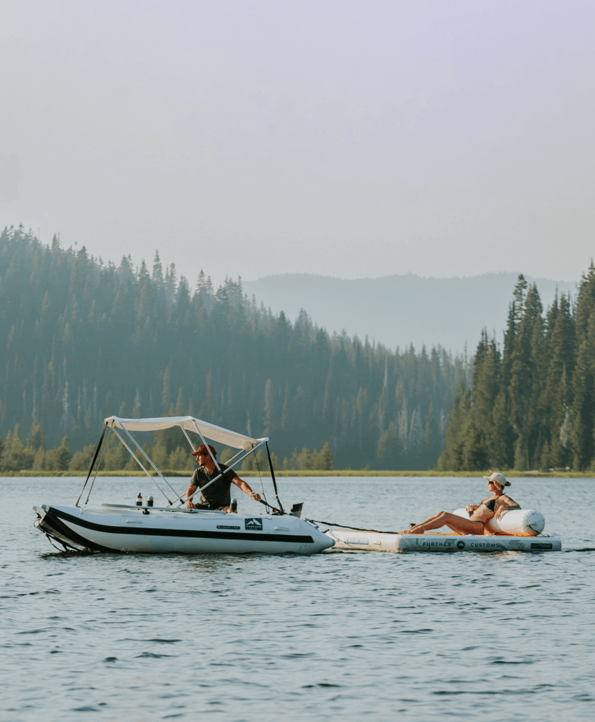 Boater towing a woman on a lounge float in a serene lake surrounded by forested hills under a clear sky.