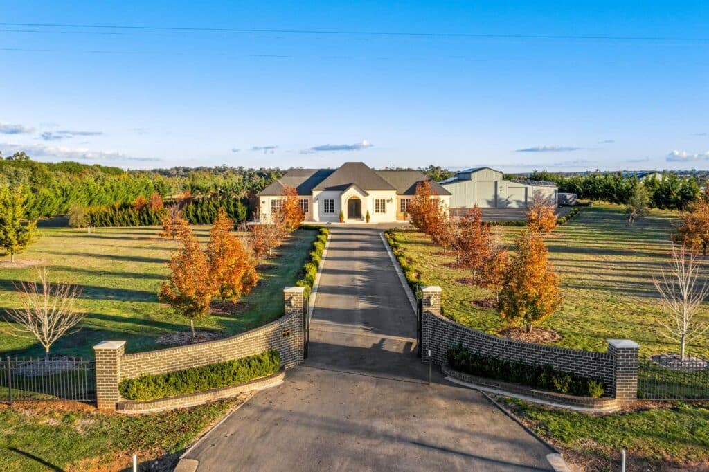 Luxurious countryside estate with tree-lined driveway, expansive gardens, and a grand entrance under a clear blue sky.