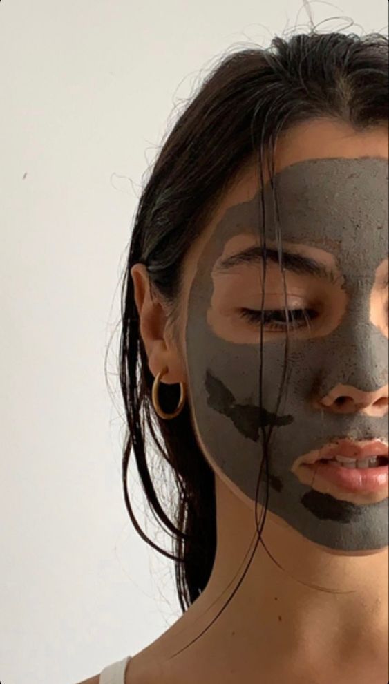 Woman with gray clay face mask and wet hair, wearing a hoop earring, against a plain background. Skincare and beauty treatment.