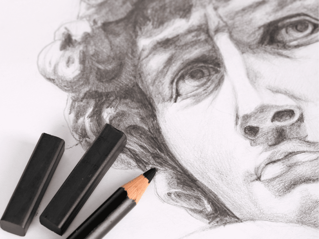Detailed pencil sketch of a classical sculpture's face with charcoal blocks and pencil nearby, highlighting artistic techniques.