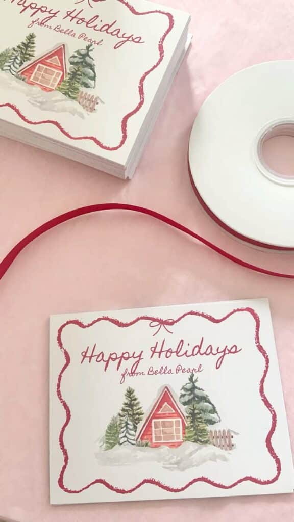 christmas business packaging 🎄 Holiday cards featuring a winter cabin design with Happy Holidays from Bella Pearl text, accompanied by red ribbon on a pink surface. | Sky Rye Design Holiday cards featuring a winter cabin design with Happy Holidays from Bella Pearl text, accompanied by red ribbon on a pink surface.