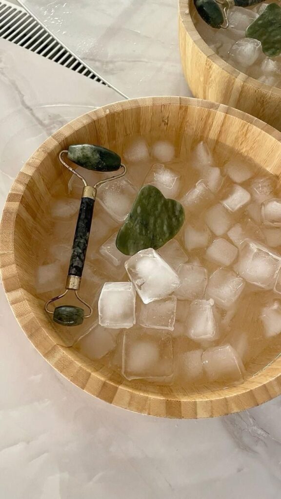 Jade roller and gua sha tool in a bamboo bowl with ice cubes, ideal for natural skincare and facial massage routines.