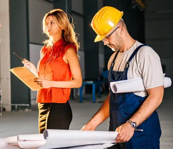 construction-estimating-services Woman with clipboard and man in hard hat reviewing blueprints at construction site. | Sky Rye Design Woman with clipboard and man in hard hat reviewing blueprints at construction site.