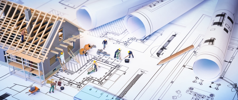 Miniature construction workers building a house on architectural blueprints, surrounded by rolled designs and a pencil, illustrating planning.