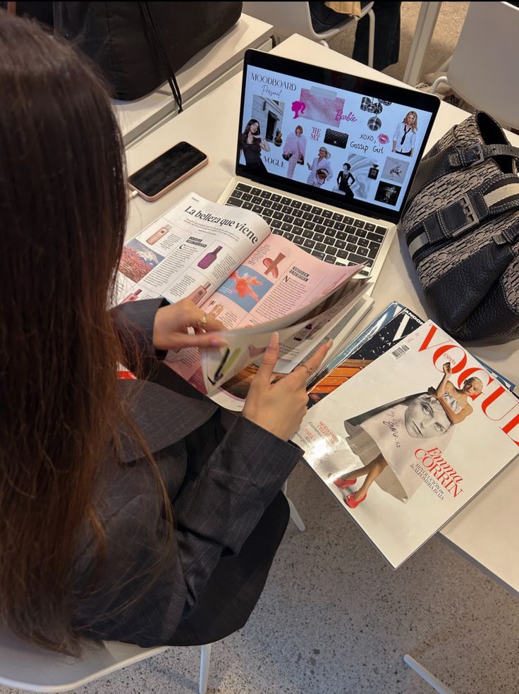 Woman reading fashion magazine at desk with laptop showing moodboard, surrounded by Vogue magazines and accessories. Fashion inspiration.