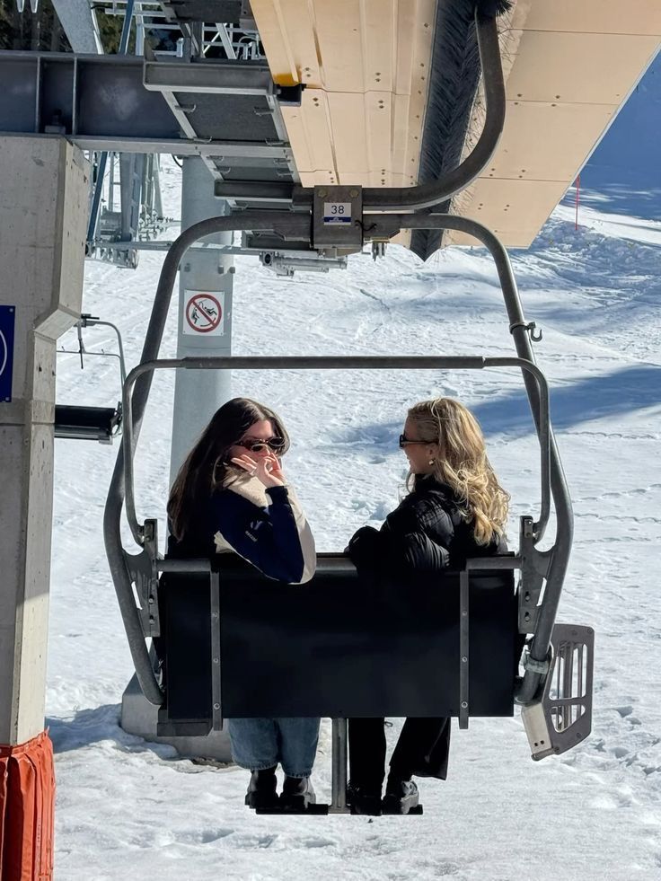 Two people on a ski lift enjoying a sunny day on the snowy slopes, wearing winter jackets and sunglasses.