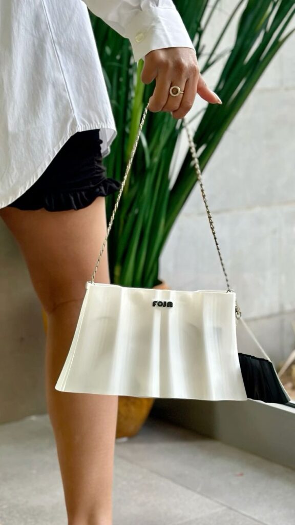 Person holding a stylish white handbag with a chain strap, wearing a white shirt and black shorts, plants in the background.