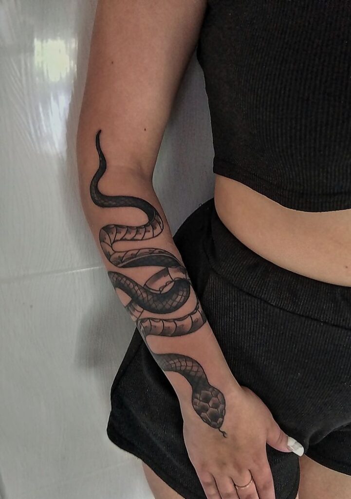 Forearm tattoo of a detailed black snake on a person wearing a black top and shorts, showcasing intricate design and texture.