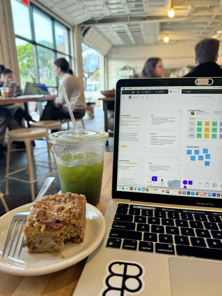 Laptop with open screen, iced matcha drink, and cake slice on a café table. People working in background through large windows.