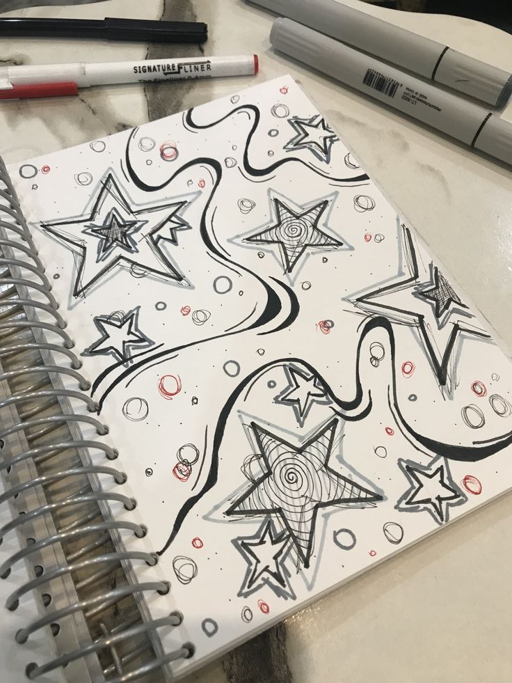 Sketchbook page with doodles of stars, swirling lines, and circles next to markers on a table, showcasing creative designs.