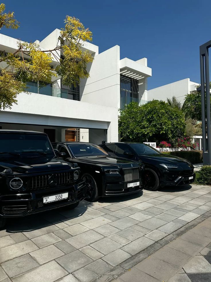 Luxury black cars parked in front of a modern white house, surrounded by greenery and clear blue skies.