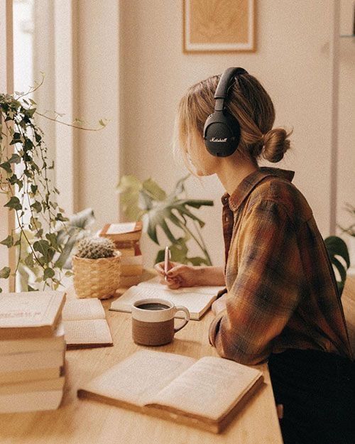e77e540eb514aa7d4787ac596ba12729 Woman writing at a desk with headphones, surrounded by books and plants, creating a cozy workspace ambiance with natural light. | Sky Rye Design Woman writing at a desk with headphones, surrounded by books and plants, creating a cozy workspace ambiance with natural light.
